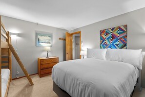 Une chambre à coucher avec un lit, une commode en bois, une table de chevet, une lampe, un tableau et une porte.
