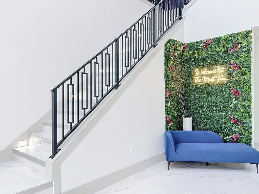 Una escalera con barandilla negra, una pared verde con plantas, un letrero de neón y un sofá azul.