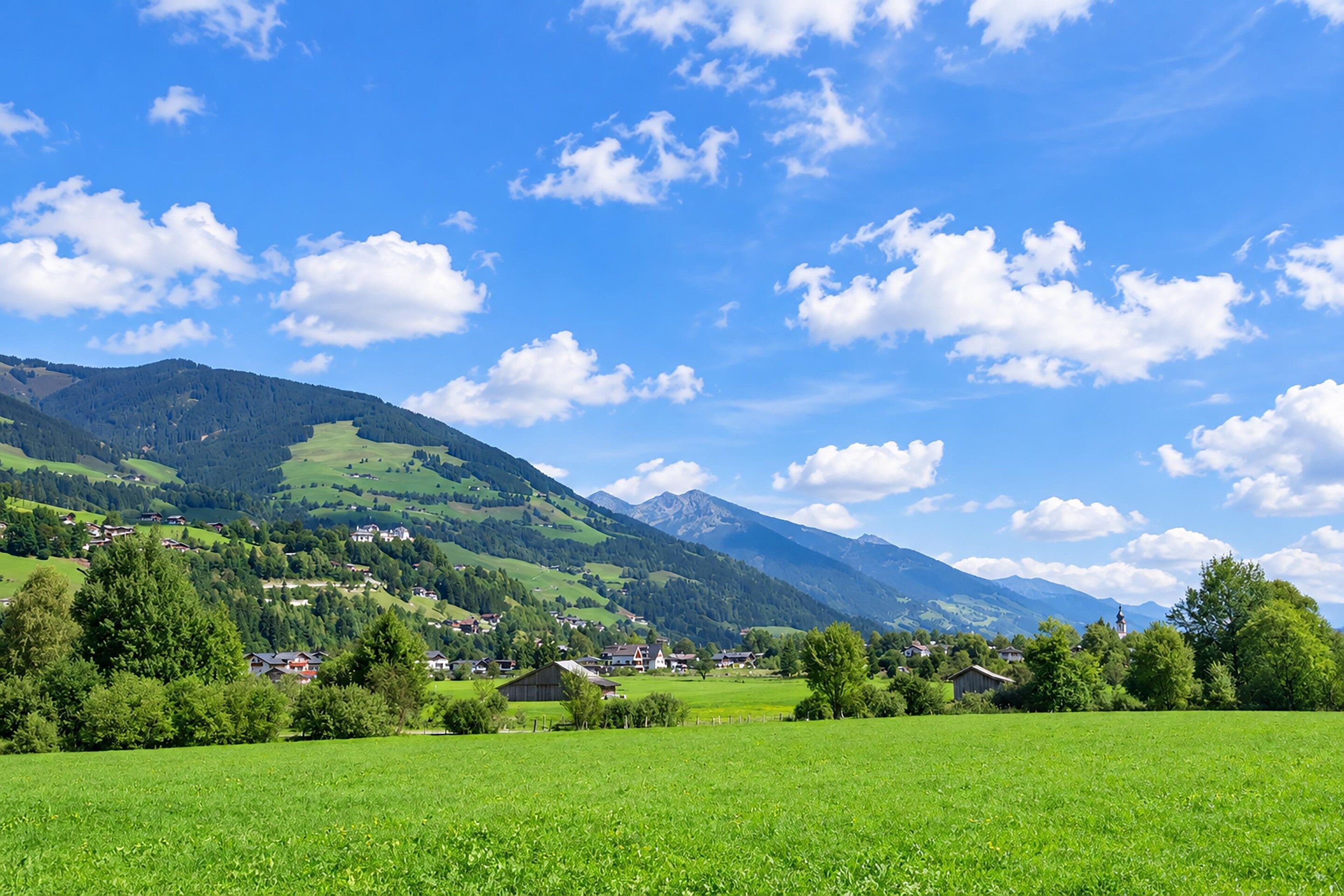 Ländliche Landschaft mit einem Dorf, eingebettet zwischen grünen Hügeln und Bergen unter blauem Himmel.
