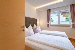 Ein Hotelzimmer mit zwei Betten, einer Holztür, einem Fenster mit Vorhängen und einem Kopfteil mit einer Leuchte.
