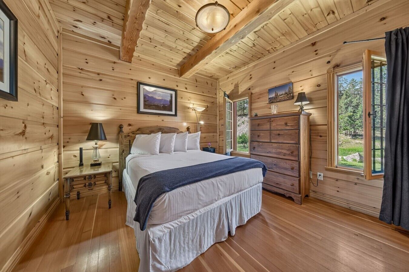 A wooden cabin bedroom with a bed, dresser, bedside table, lamp, and framed pictures.
