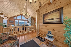 Ein Interieur einer Holzhütte mit Kamin aus Stein, einer hölzernen Bank, einer Couch, einem Esstisch und Blick auf Berge durch große Fenster.