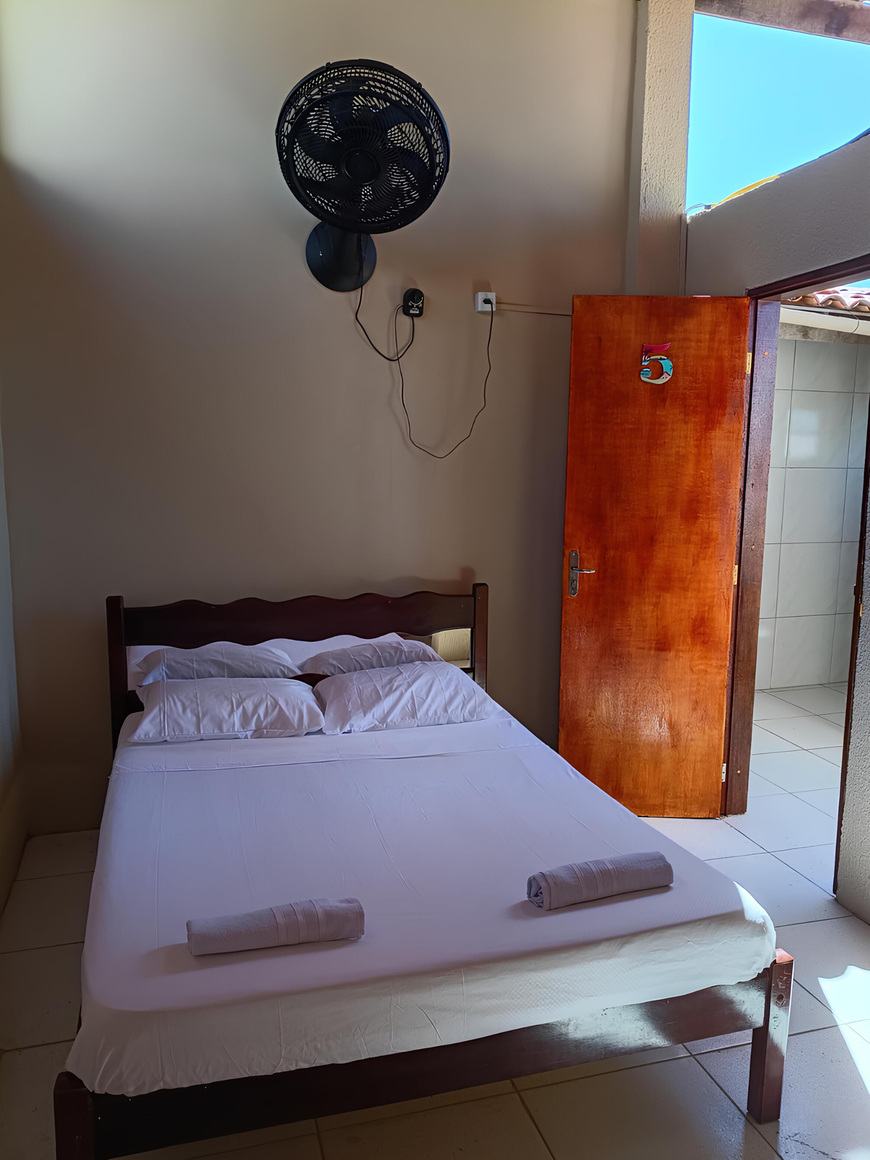 A bed with white linens, a wall-mounted fan, and a wooden door.