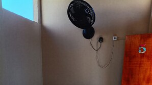 Una cama individual con ropa de cama blanca y dos toallas enrolladas, un ventilador de pared y una puerta de madera.