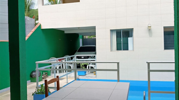 Un espacio al aire libre techado con una mesa y sillas, una piscina y un edificio con paredes blancas y detalles en verde.