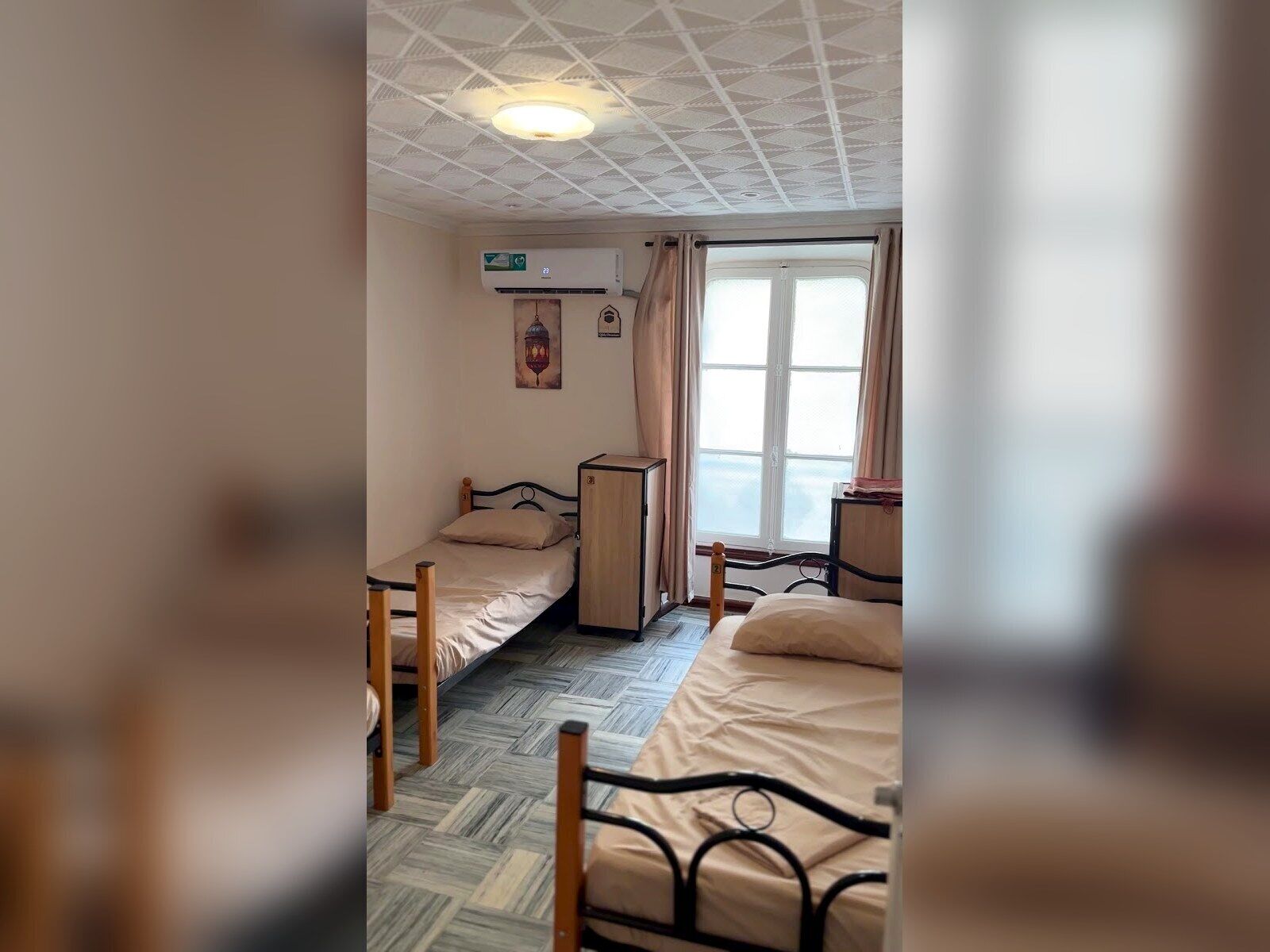 Quarto com duas camas, uma janela, um ventilador de teto e um aparelho de ar condicionado.