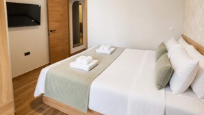 Un lit bien fait, avec du linge de lit blanc et un plaid vert, une porte en bois et un téléviseur fixé au mur.