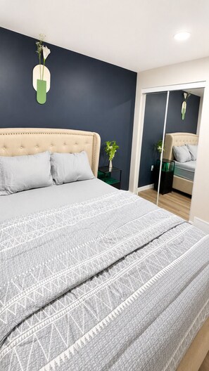 A bedroom with a large bed, a mirror, and a green vase with flowers on the wall. - Your Devonshire, Transcona Retreat | Cozy 2BR Haven (Winnipeg)
