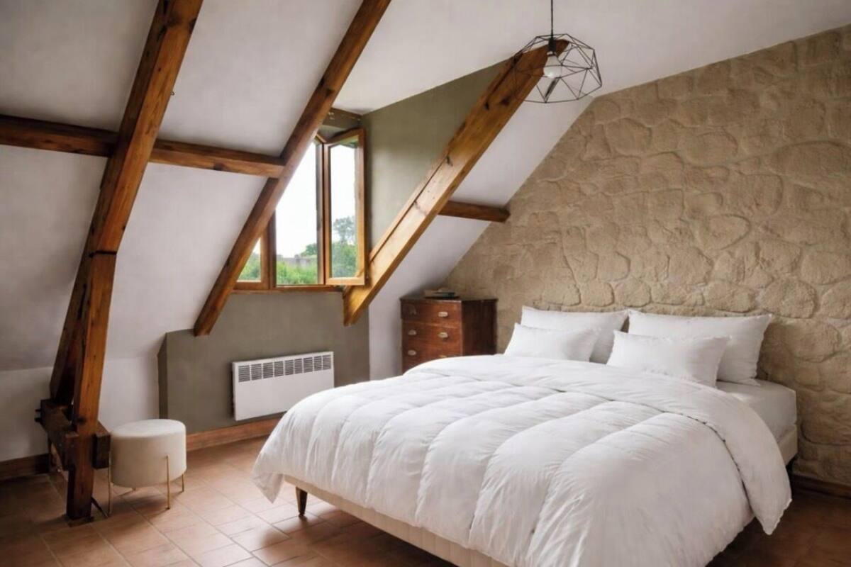 Un dormitorio con techo inclinado, pared de piedra, cama con ropa blanca, una silla de madera y una ventana.