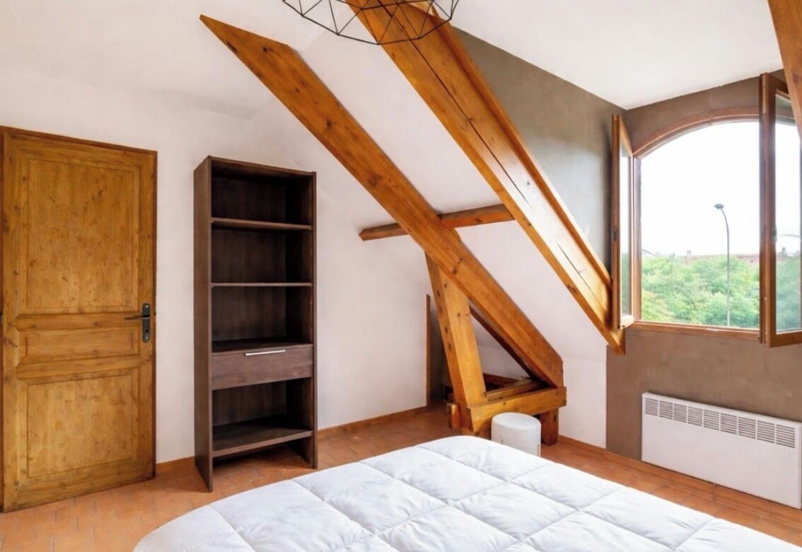 A bedroom with a sloped ceiling, a wooden door, a built-in shelf, a window, and a radiator.