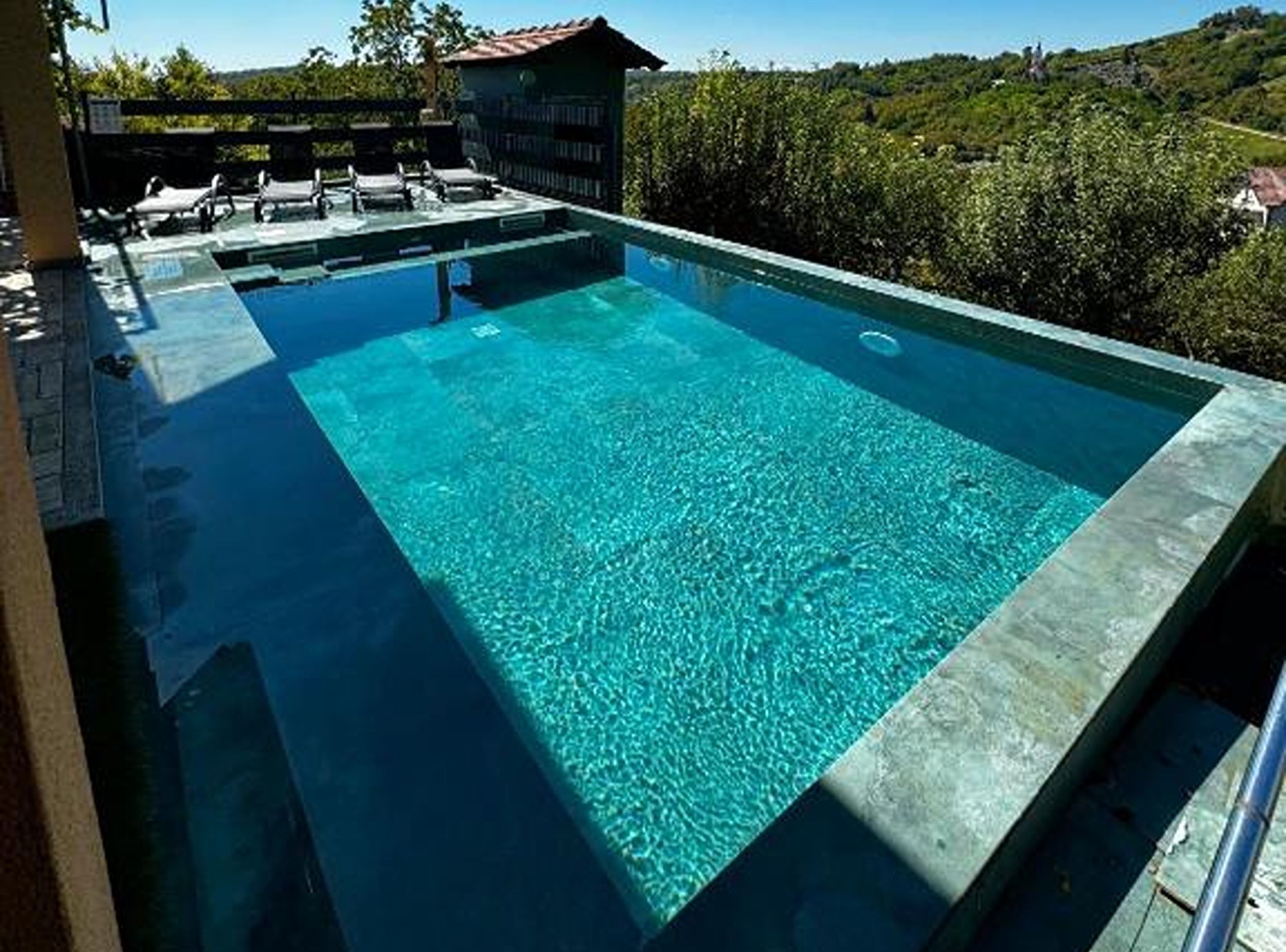 A rectangular swimming pool with clear blue water, surrounded by a stone deck and lounge chairs.