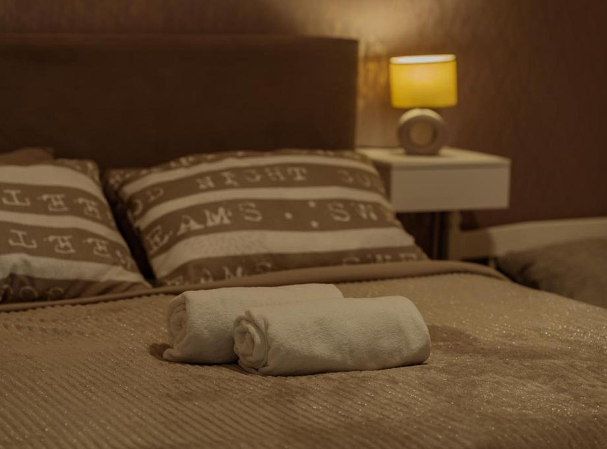 A neatly made bed with rolled towels, a headboard, and a bedside lamp.