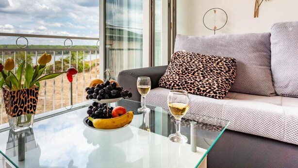 A balcony with a glass table, a sofa with leopard print cushions, a bowl of fruit, and a vase with flowers.