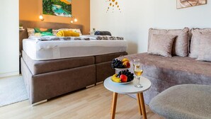 A bedroom with a bed, a sofa, a small table with fruit and a glass of wine, and a painting on the wall.
