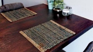 Comedor con mesa de madera, dos individuales tejidos, un reloj y una planta pequeña con un candelabro.