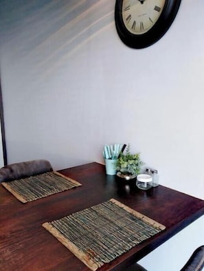 A dining area with a wooden table, two woven placemats, a clock, and a small plant with a candle holder.