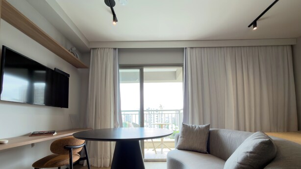 Ein modernes Wohnzimmer mit Sofa, rundem Tisch, an der Wand montiertem Fernseher und einem Balkon mit Vorhängen.