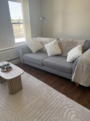 Ein Wohnzimmer mit grauem Sofa, weißen gemusterten Kissen, einem runden Holztisch und einem weißen Teppich.