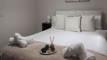 Ein ordentlich bezogenes Bett mit weißen Leinen und einer beigen Tagesdecke, ein Nachttisch mit Lampe sowie ein Tablett mit Kerze und Reed-Diffuser.