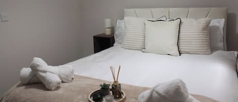 A neatly made bed with white linens and a beige throw blanket, a bedside table with a lamp, and a tray with a candle and reed diffuser.