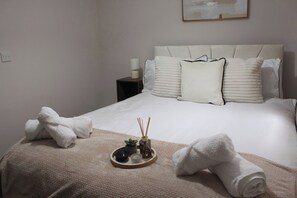 Ein ordentlich bezogenes Bett mit weißen Leinen und einer beigen Tagesdecke, ein Nachttisch mit Lampe sowie ein Tablett mit Kerze und Reed-Diffuser.
