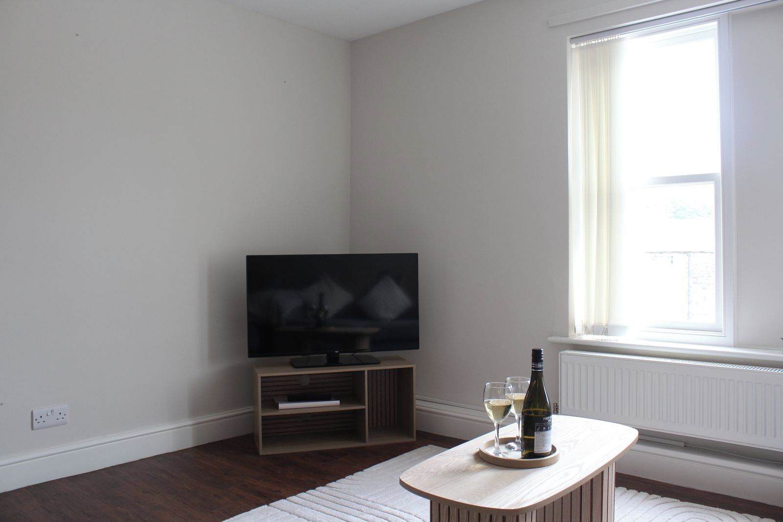 Ein Zimmer mit einem Flachbildfernseher auf einem Holzboden, einem kleinen Tisch mit einer Flasche und Gläsern sowie einem großen Fenster mit Vorhängen.