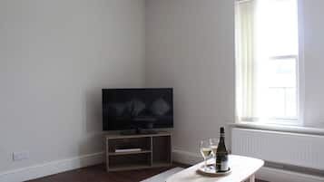 Ein Zimmer mit einem Flachbildfernseher auf einem Holzboden, einem kleinen Tisch mit einer Flasche und Gläsern sowie einem großen Fenster mit Vorhängen.