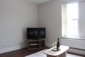 Ein Zimmer mit einem Flachbildfernseher auf einem Holzboden, einem kleinen Tisch mit einer Flasche und Gläsern sowie einem großen Fenster mit Vorhängen.