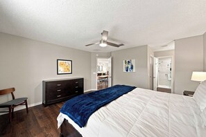 Une chambre à coucher comprenant un lit, une commode, une chaise, un ventilateur de plafond et une porte donnant sur une salle de bain.
