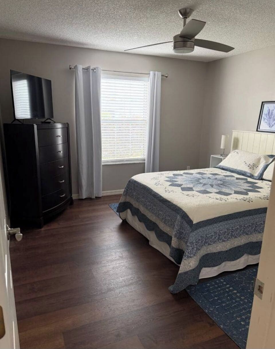 Um quarto com cama, ventoinha no teto, cômoda e janela com cortinas.
