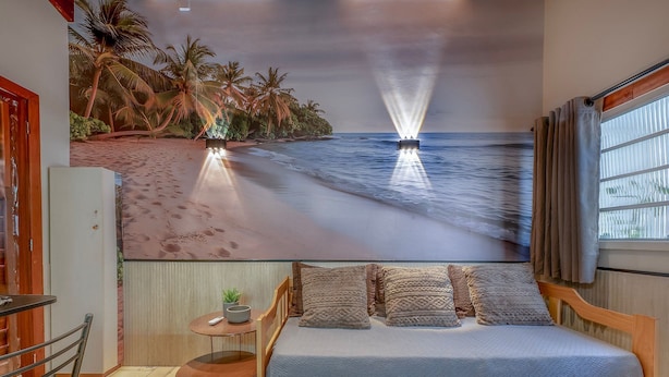 Een slaapkamer met een bed, een klein tafeltje met een plant, een stoel en een muurschildering van een strand.