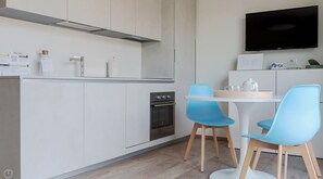 Une cuisine moderne avec des meubles blancs, un four encastré, un coin repas avec une table blanche et deux chaises bleues, et un téléviseur à écran plat fixé au mur.