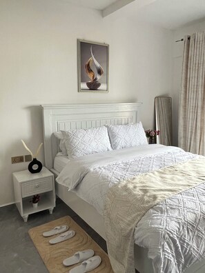 Ein Schlafzimmer mit einem weißen Bett, einem Nachttisch mit einer Vase und einem dekorativen Gegenstand, einem Spiegel und einem Paar Hausschuhen auf einem Teppich.