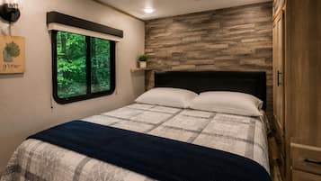 Une chambre avec un lit, une fenêtre donnant sur un paysage verdoyant, une étagère murale agrémentée d’une plante et une armoire en bois.