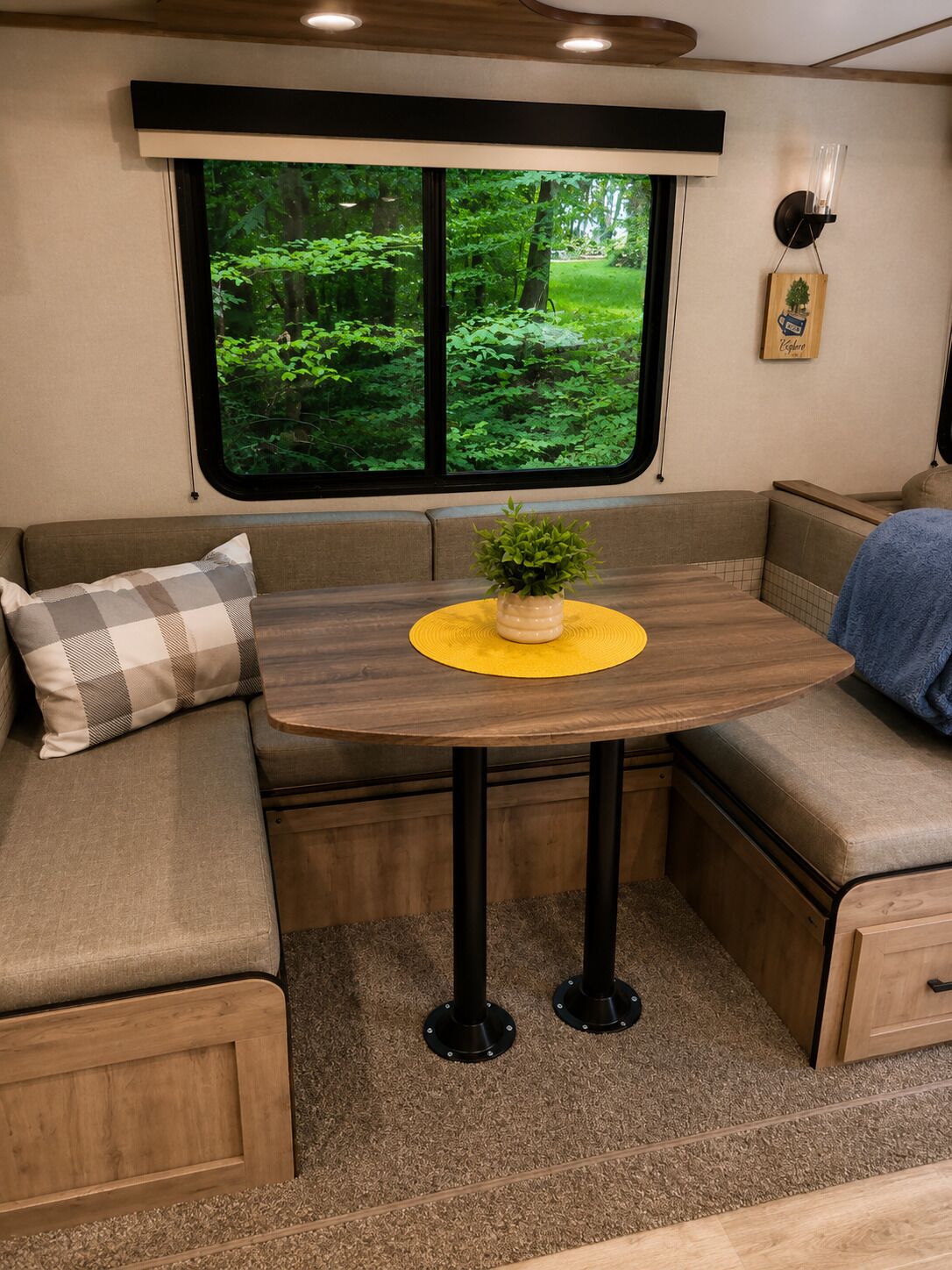 Un intérieur de camping-car accueillant, avec une table à manger, des sièges rembourrés, une fenêtre donnant sur la forêt et un luminaire fixé au mur.