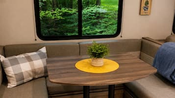 Un intérieur de camping-car accueillant, avec une table à manger, des sièges rembourrés, une fenêtre donnant sur la forêt et un luminaire fixé au mur.