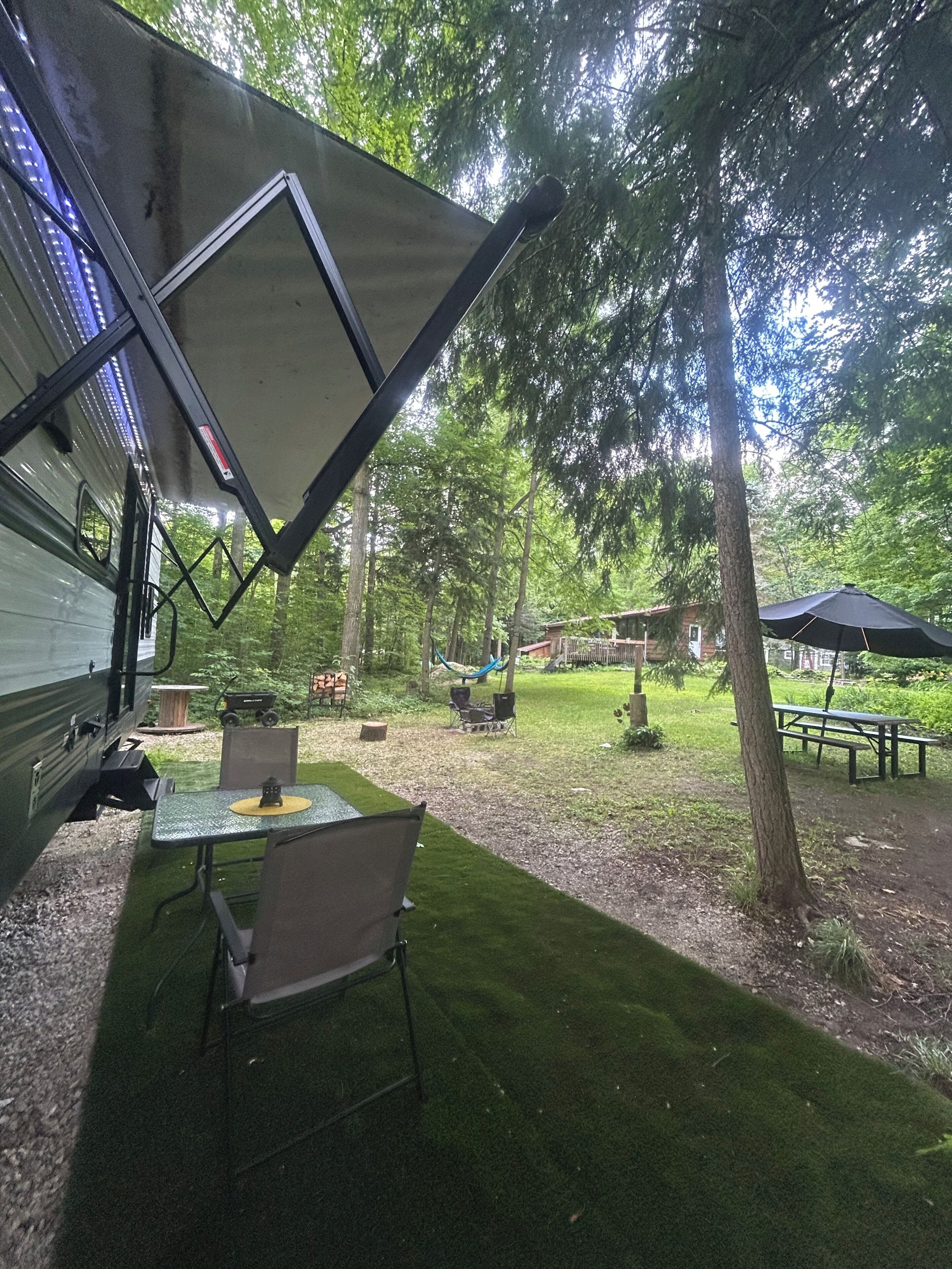 A campsite with a covered dining area, a table, and chairs, surrounded by trees and a picnic table.