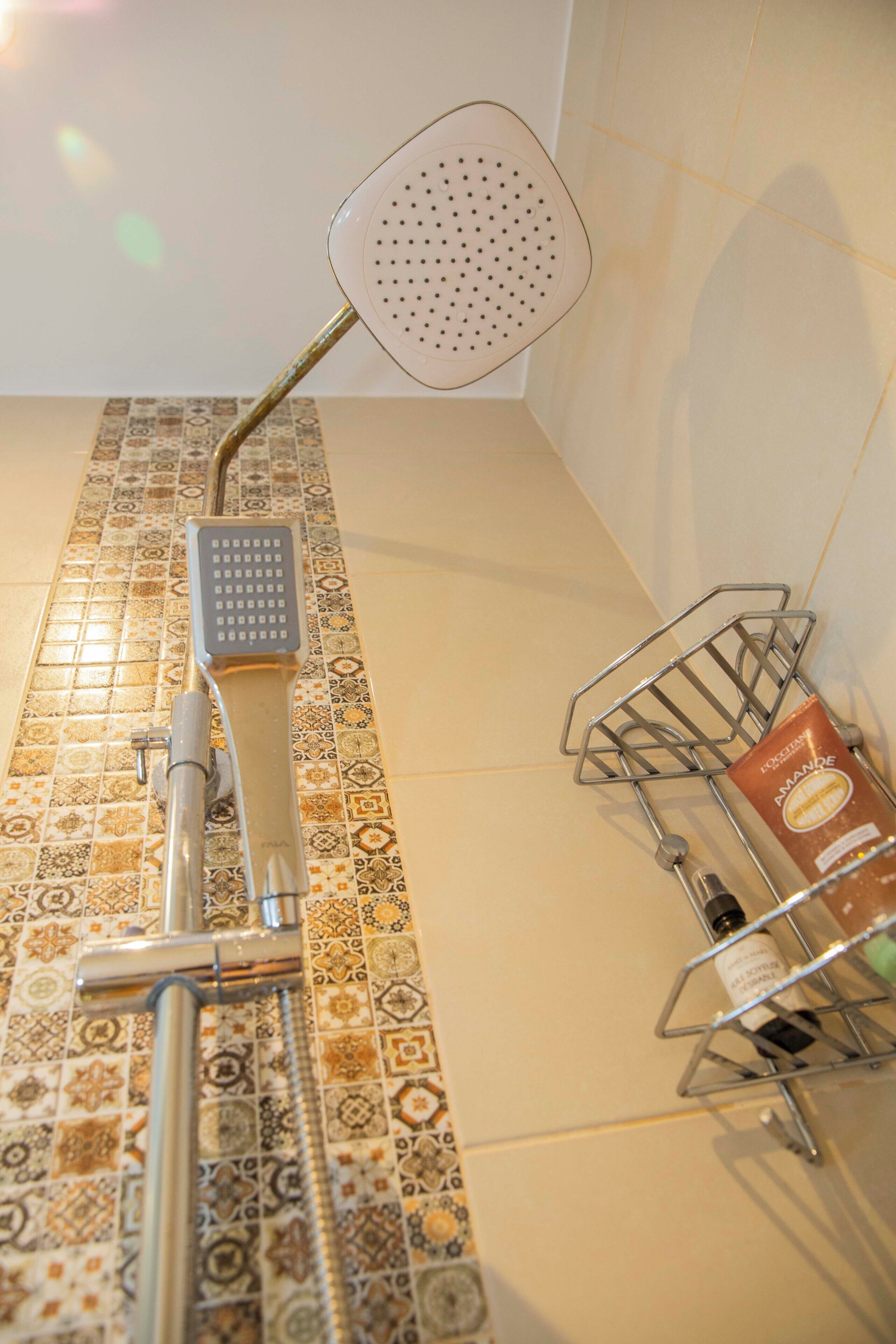 Una doccia con soffione a mano e soffione a pioggia a parete, un portaraccogli oggetti in metallo con articoli da toilette e un pavimento piastrellato con motivo geometrico.