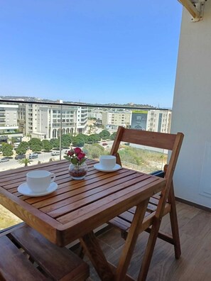 Ein Holztisch mit zwei weißen Tassen und einem Blumenstrauß auf einem Balkon mit Blick auf eine Stadtlandschaft.