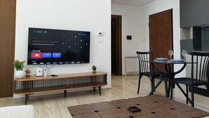 A modern living room with a flat-screen TV mounted on the wall, a wooden console table, a dining area with a table and chairs, and a kitchen area with cabinets. - Dream Apartment in Jardins de Carthage (Résidence luxoria)