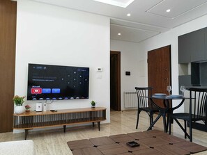 Ein modernes Wohnzimmer mit Flachbildfernseher an der Wand, einem Holzkonsolentisch, einem Essbereich mit Tisch und Stühlen sowie einer Küchenzeile mit Schränken.