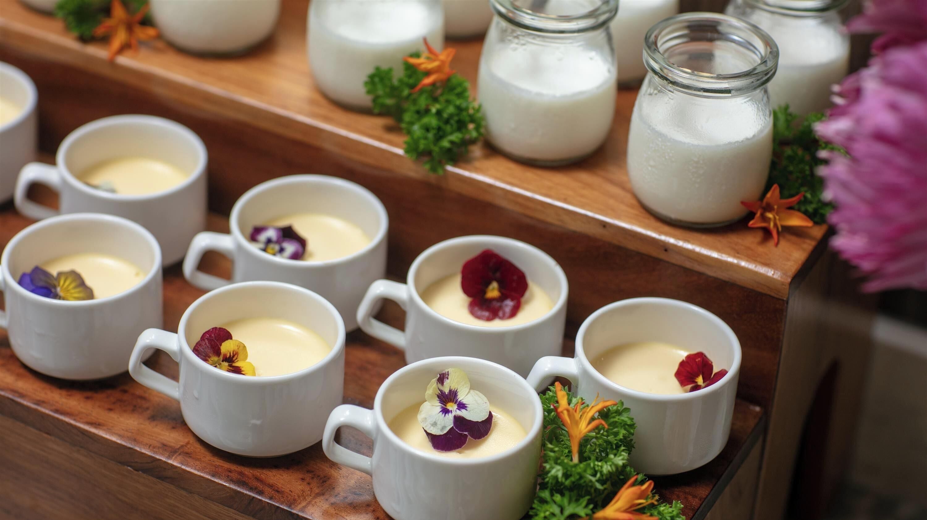 A display of white ceramic mugs with a creamy beverage, some adorned with flowers, on a wooden surface with milk bottles in the background.