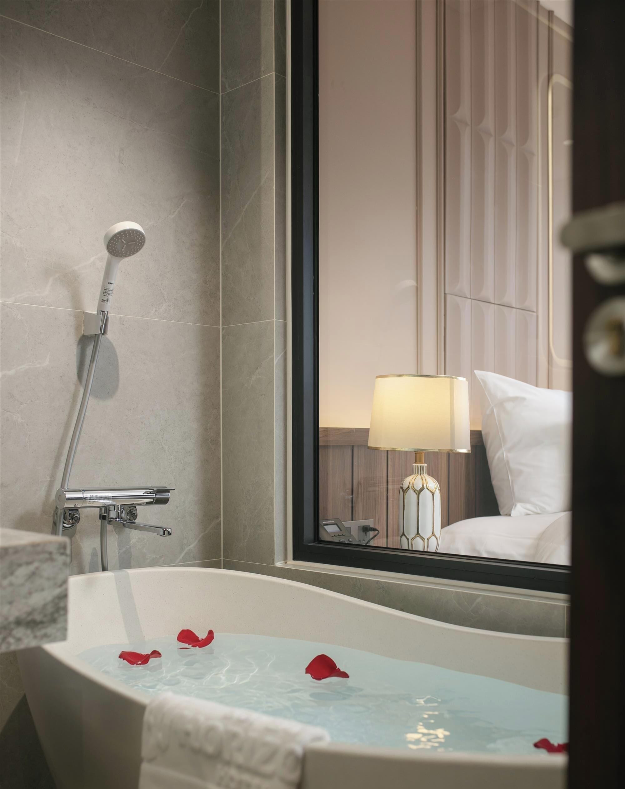 A bathroom with a bathtub, a shower, and a mirror reflecting a room with a bed and a lamp.