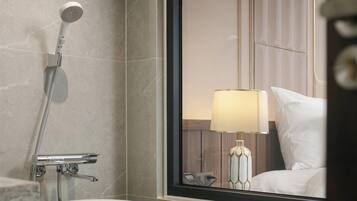 A bathroom with a bathtub, a shower, and a mirror reflecting a room with a bed and a lamp.