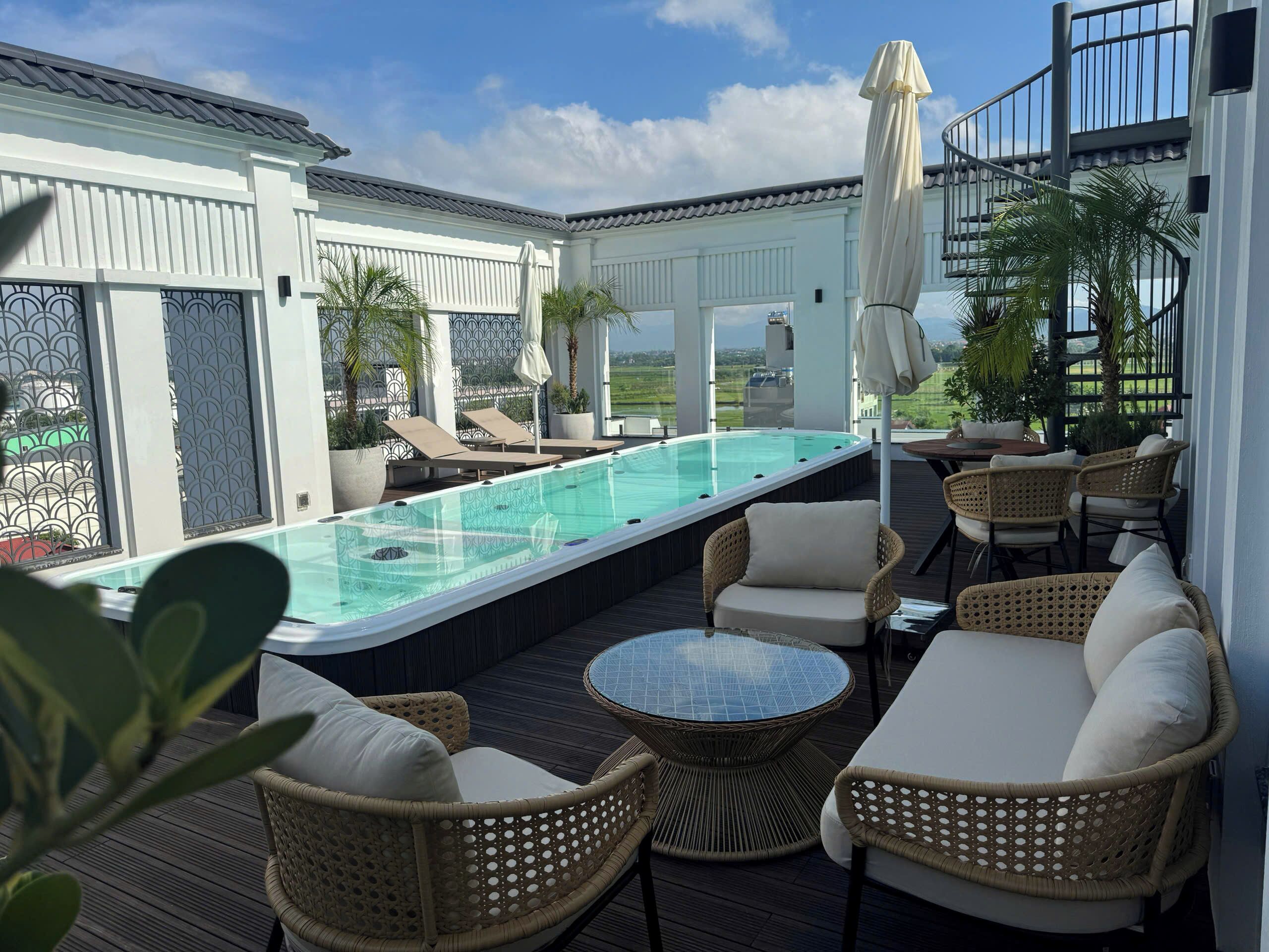 An outdoor pool area with lounge chairs and a table, surrounded by a white building with a tiled roof.