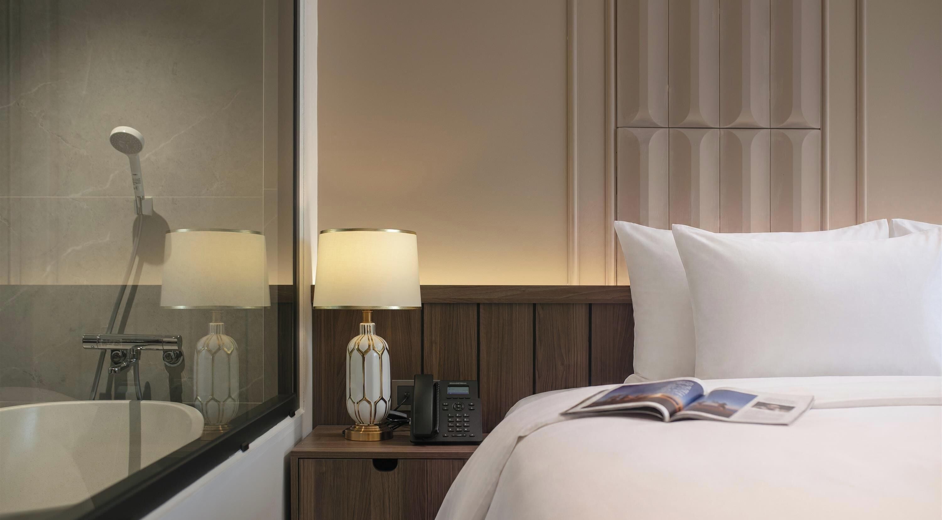 A hotel room with a bed, a nightstand with a lamp and phone, a mirror, and a shower.