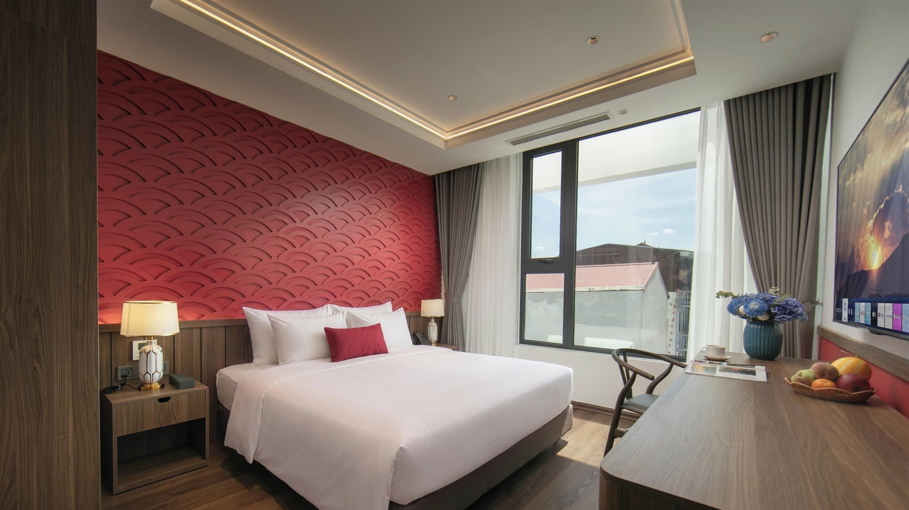 Ein Hotelzimmer mit einem großen Bett, einem Schreibtisch mit Stuhl, einem Fenster mit Aussicht und einer roten Wand mit Struktur.
