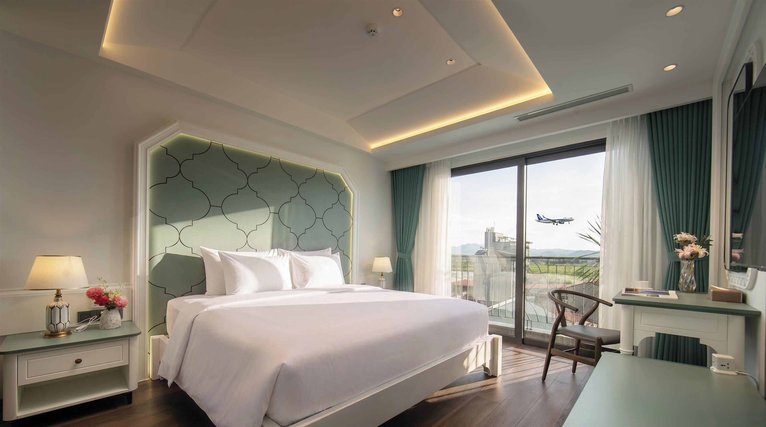 A hotel room with a large bed, a desk with a chair, a view of the outdoors, and a modern ceiling design.