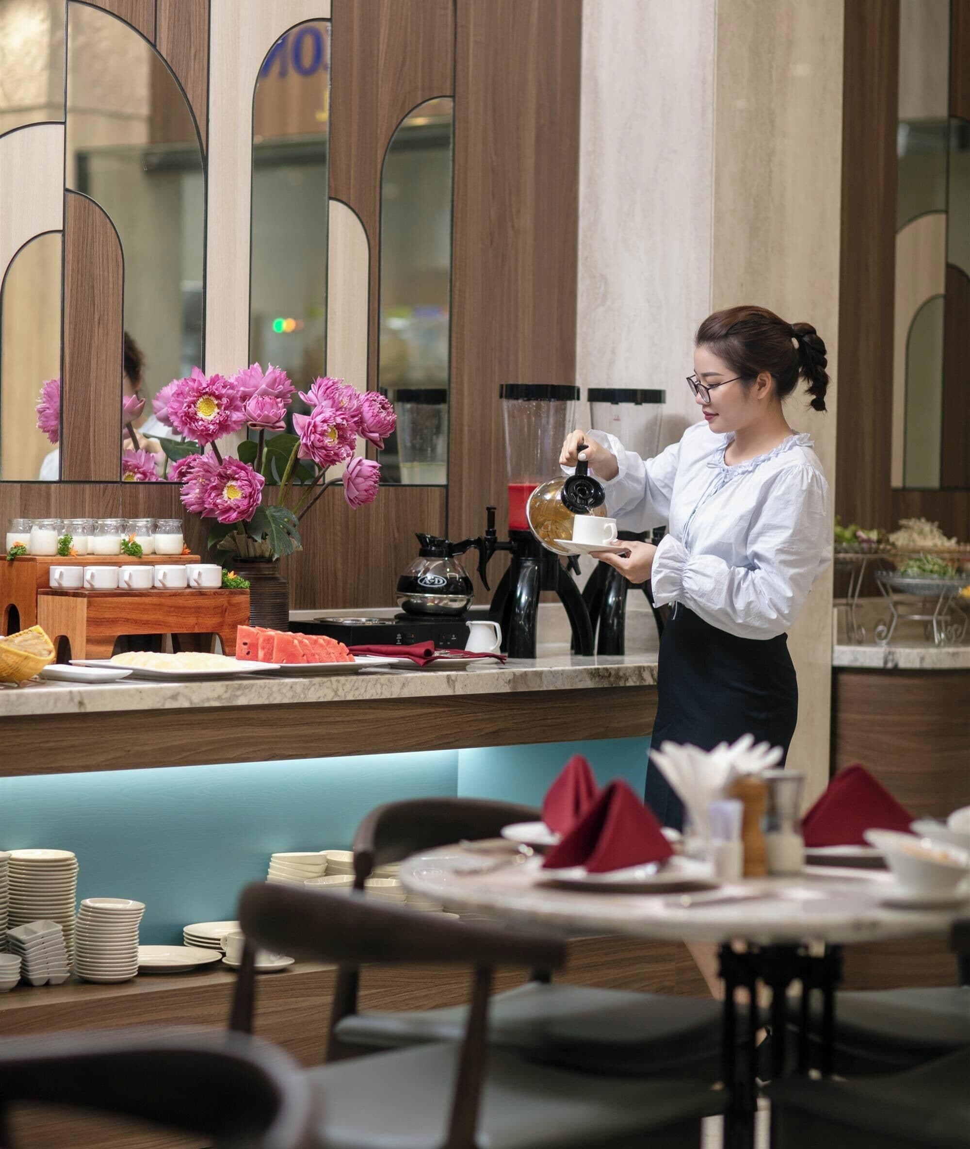 A person serving coffee in a restaurant with a counter, a flower arrangement, and neatly arranged tables and chairs.
