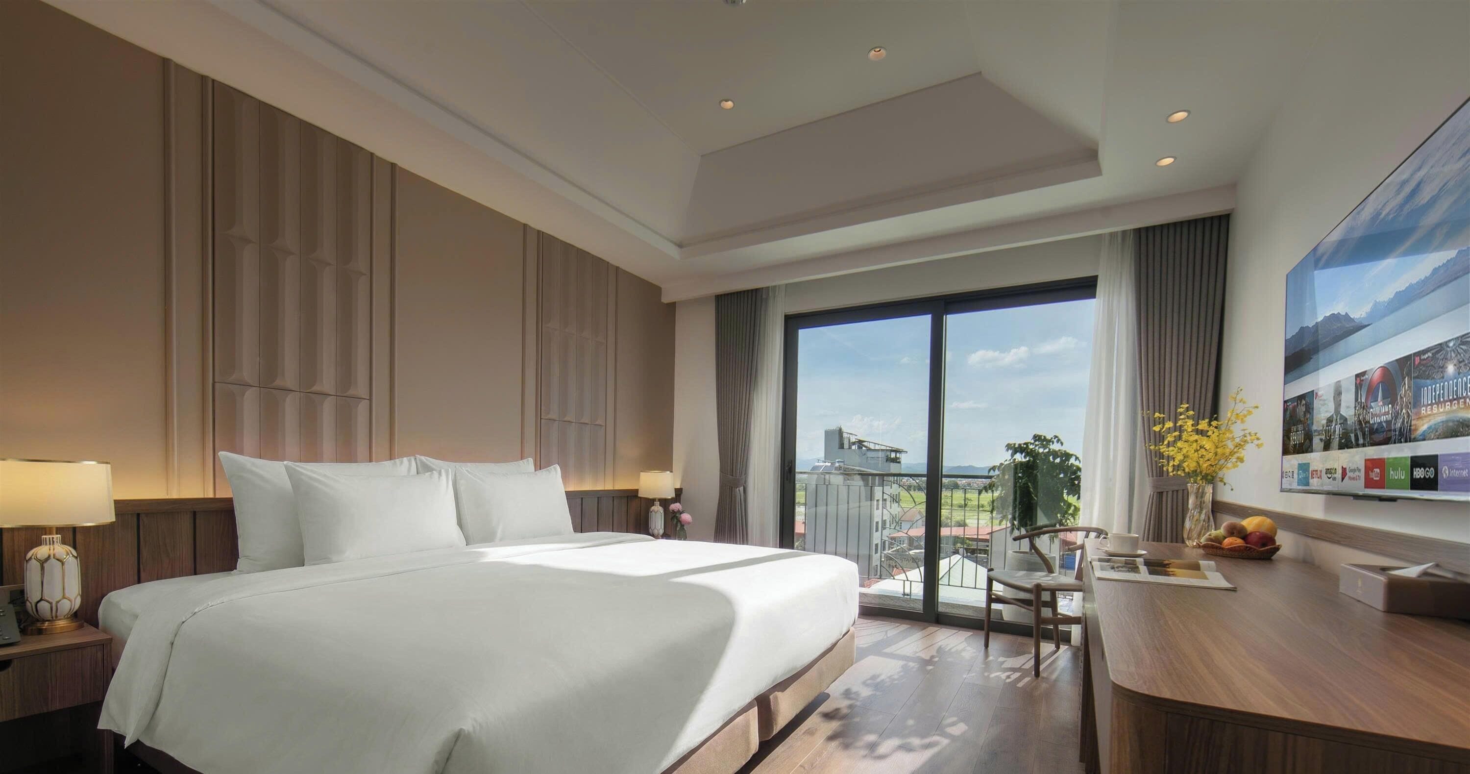 A hotel room with a large bed, a desk with a chair, a view through a large window, and a wall-mounted TV.